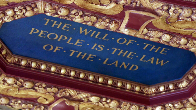 A fresco with the painted words THE • WILL • OF • THE • PEOPLE • IS • THE • LAW • OF • THE • LAND is framed by painted carved wood in flower, leaf and fruit motifs.