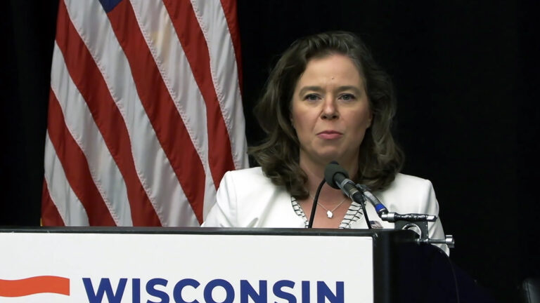 Sara Rodriguez speaks into multiple microphones mounted to the top of a podium with a poster with the word Wisconsin affixed to its front while standing in front of a U.S. flag.