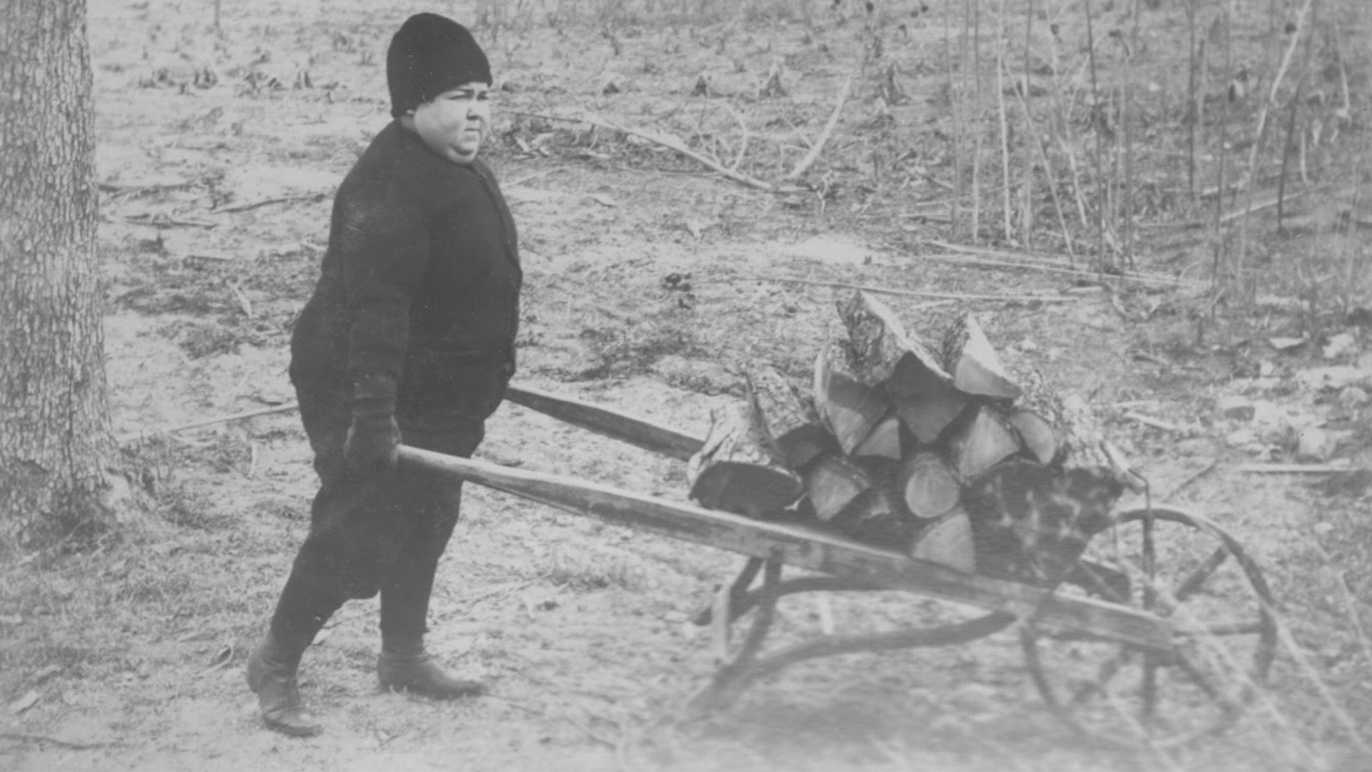 Lloyd "Babe" Spencer as a young child, dressed warmly in a hat and coat, pushes a wooden wheelbarrow filled with chopped logs through a barren, wintry landscape.