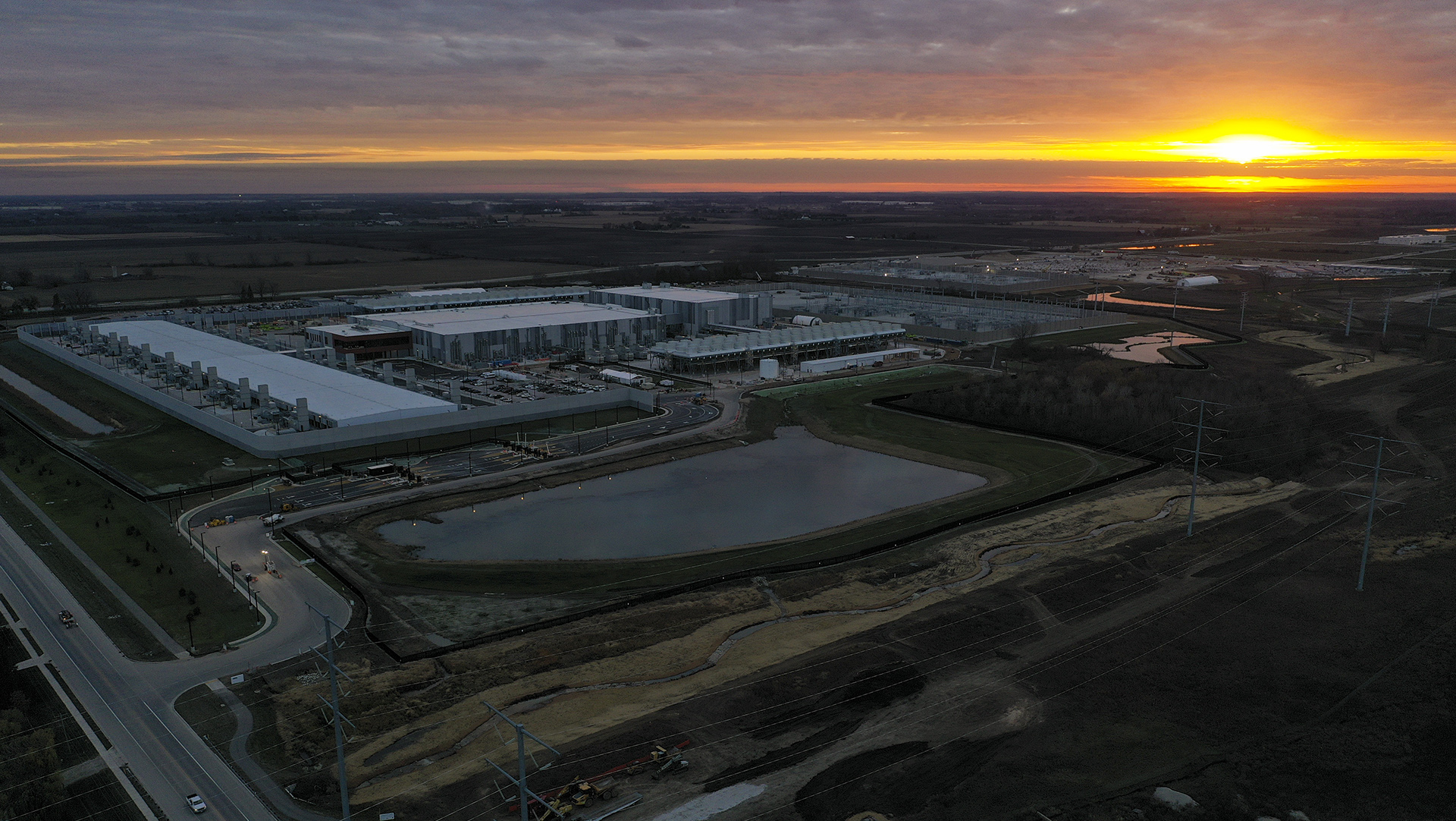 The sun sets in a partly cloudy sky over a landscape of multiple low, large buildings surrounded by roads, retention ponds, power line poles and fields.