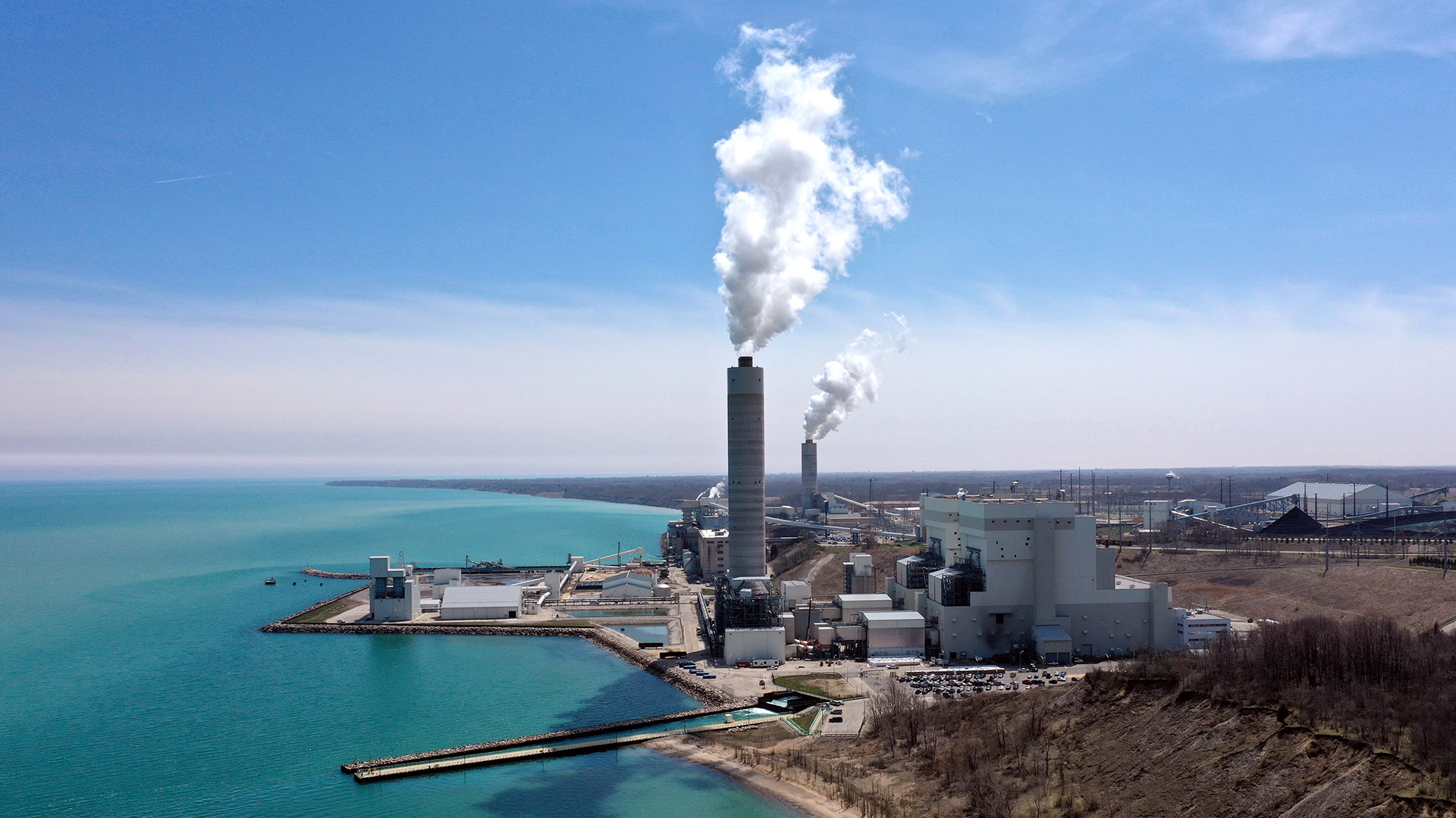 Two gas-emitting stacks stand among a collection of buildings on the shore of a large body of water, with a wooded bluff in the foreground on land and more buildings, water towers and power line poles in the background.