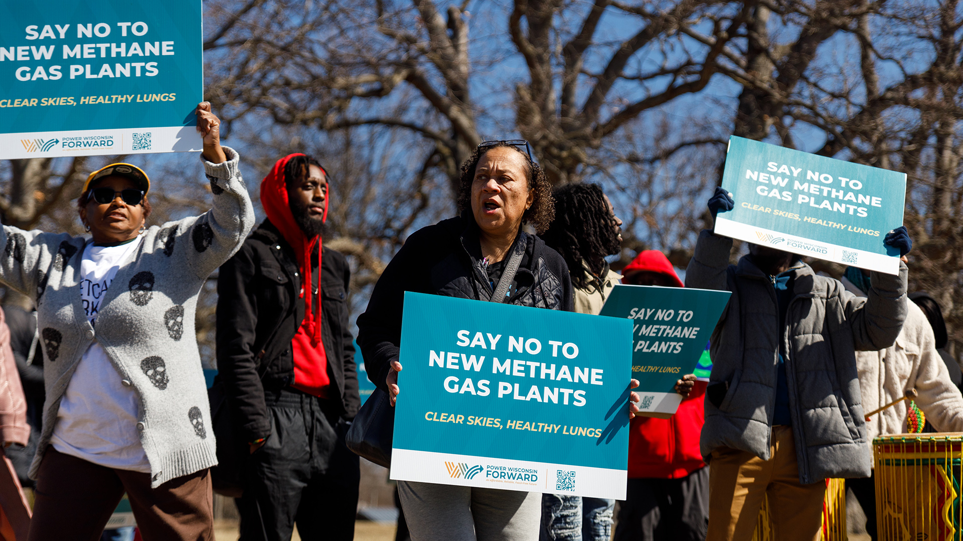 Multiple protestors stand with several holding printed signs with the words "Say No to New Methane Gas Plants" and "Clear Skies, Healthy Lungs," with leafless trees in the background.