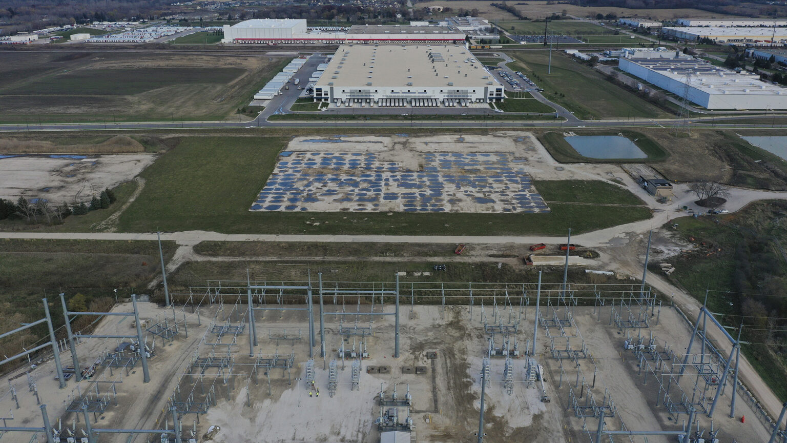An aerial photo shows an electrical substation, a concrete pad marking the location of a former building, and multiple new buildings standing among fields, retention ponds and powerline towers, with fields and wooded areas in the background.