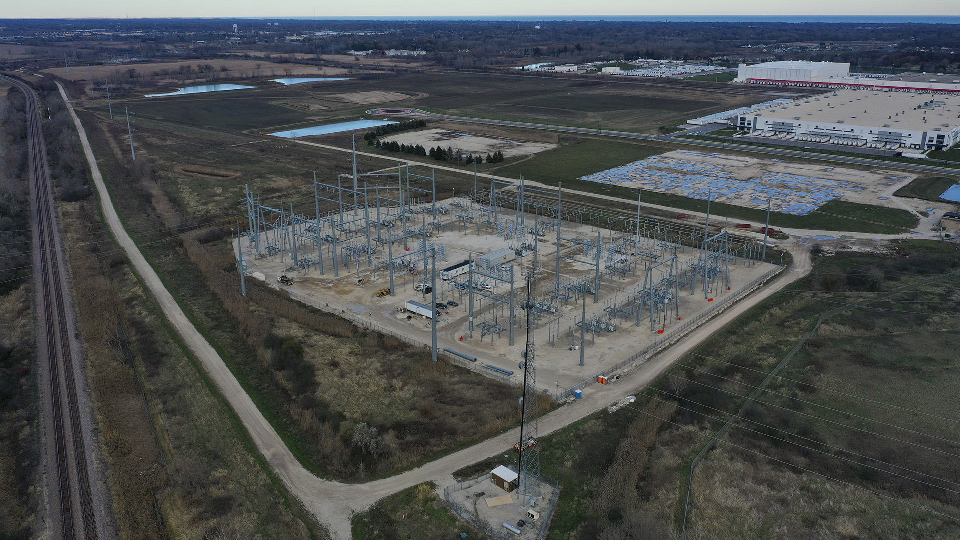 An aerial photo shows a rail line and parallel gravel road next to an electrical substation, a concrete pad marking the location of a former building, and multiple new buildings standing among fields, retention ponds and powerline towers, with fields, wooded areas and a large body of water stretching to the horizon.