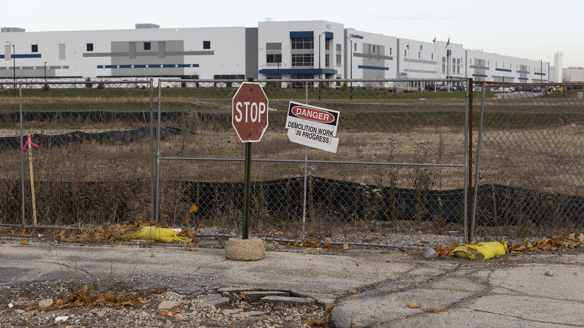 A stop sign connected by a metal rod to a concrete base stands next to a chain-link fence with a sign reading "Danger" and "Demolition Work in Progress" affixed to its side, with an empty field and large three-story building with an adjacent parking lot in the background.