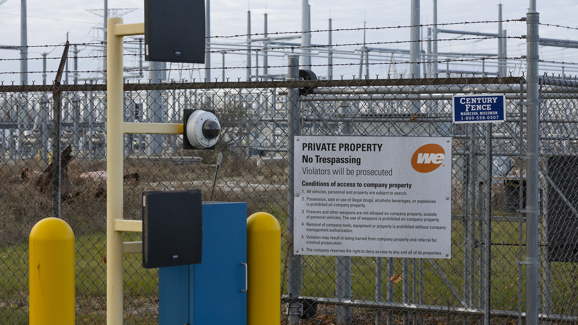 A half-sphere video camera and two electronic devices are mounted to multiple arms on a metal poll next to a metal cabinet and between two bollards next to a chain-link fence topped with barbed wire and a sign reading "Private Property," "No Trespassing," Violators will be prosecuted" and other text, wit out-of-focus towers and wires of an electrical substation in the background.