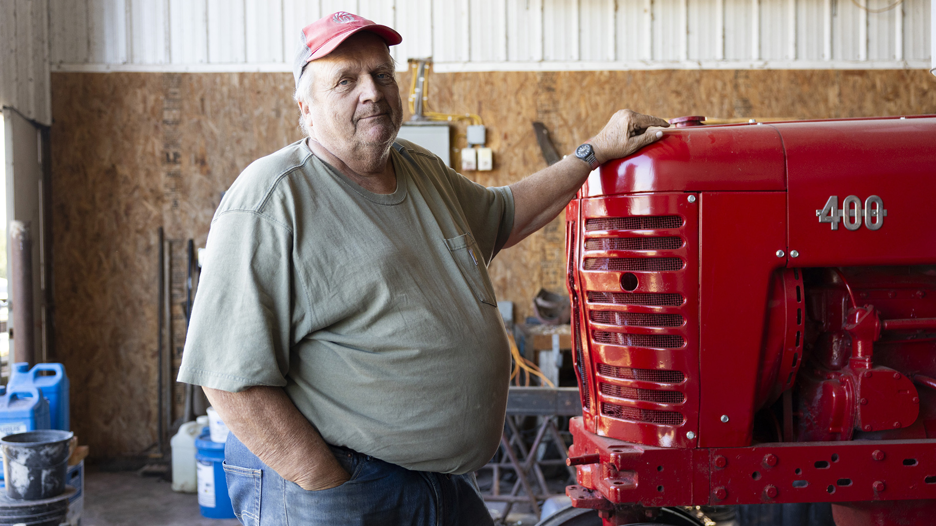 David Hardtke poses for a portrait while standing with his left hand on the hood of a farm vehicle inside a barn with plywood and metal walls.