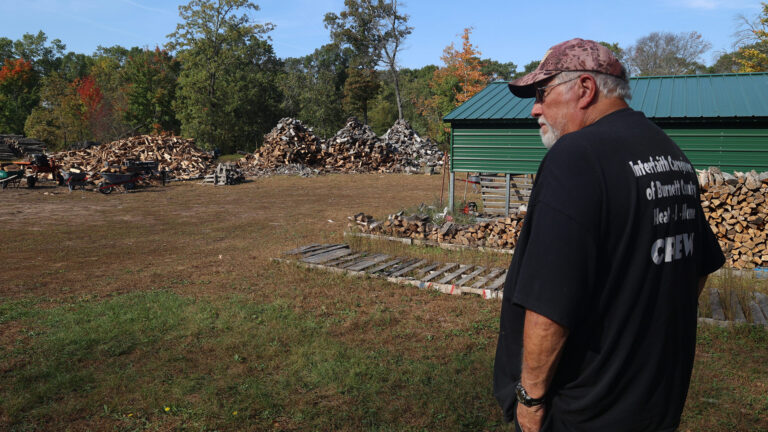Denny Blodgett stands in front of a a metal shed with stacks of split firewood along its side, with multiple piles of split firewood at the far end of a lawn in front of a line of trees with leaves of multiple colors. Denny Blodgett stands in front of a a metal shed with stacks of split firewood along its side, with multiple piles of split firewood at the far end of a lawn in front of a line of trees with leaves of multiple colors.