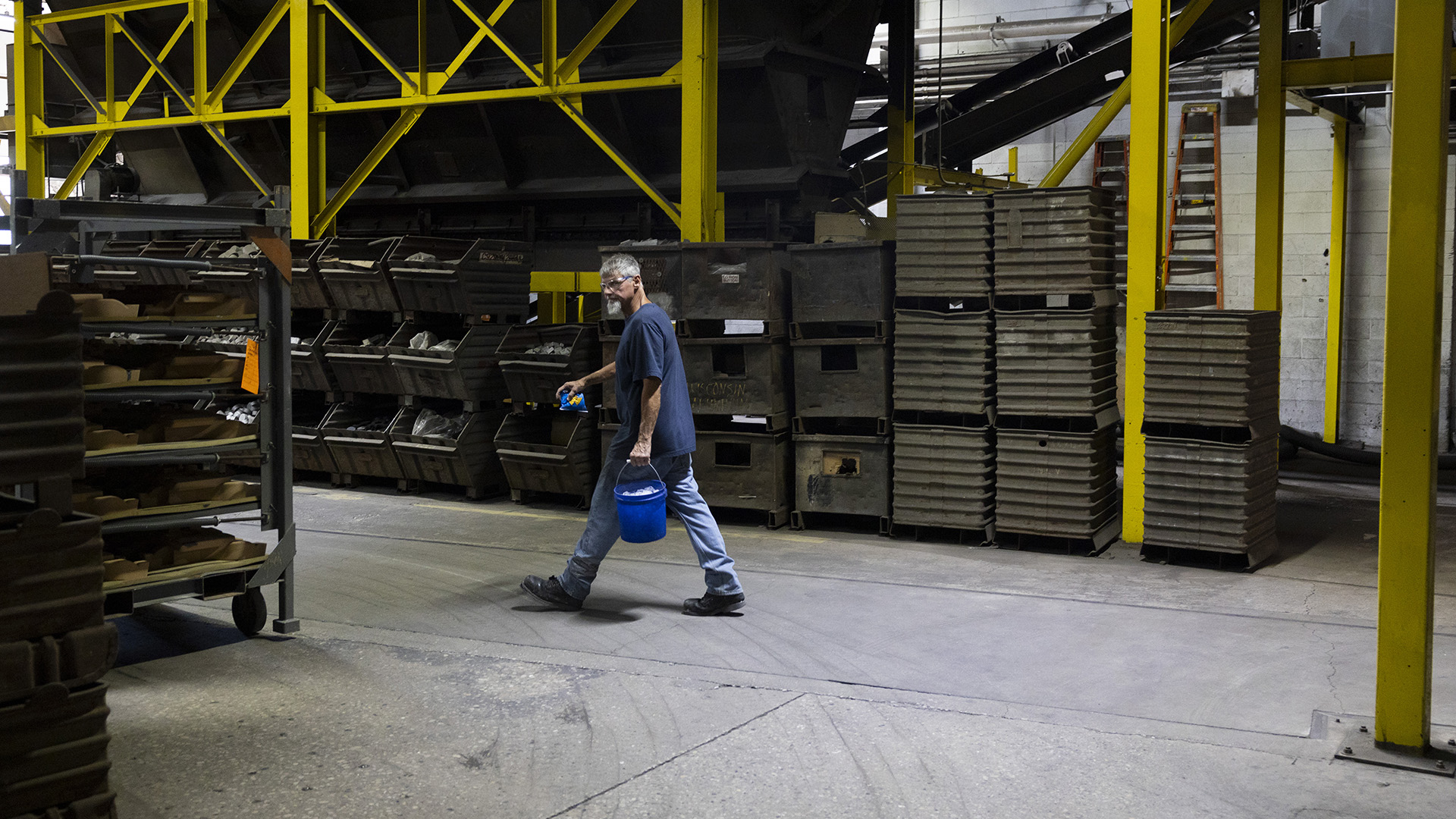 A man carrying a plastic bucket in his left hand walks on a concrete floor between a rolling metal shelving cart and stacks of storage bins in a room with a concrete floor and metal pillars.
