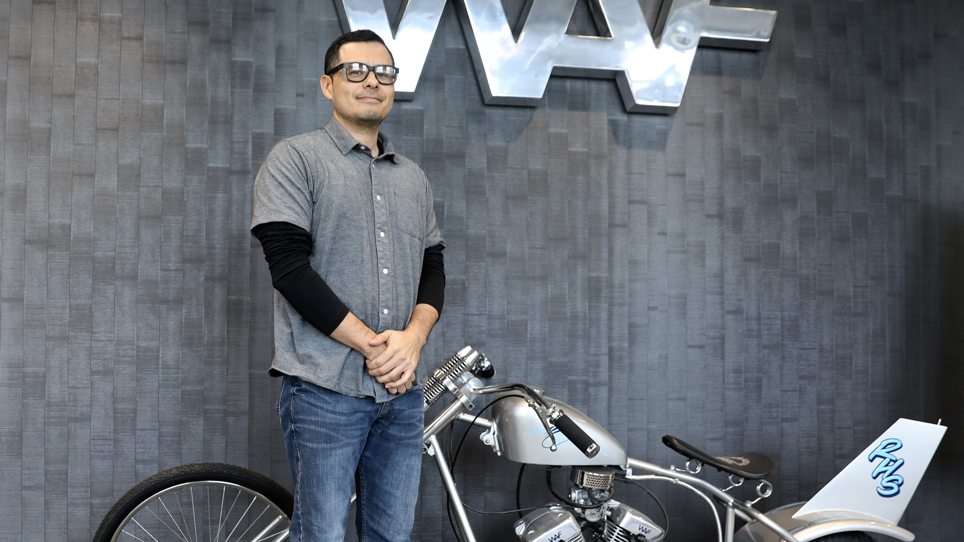 Ricardo Manriquez poses for a portrait while standing in front of a motorcycle parked in front of a wall with a metal wordmark sign with the letters "WAF" mounted to a wall in the background.
