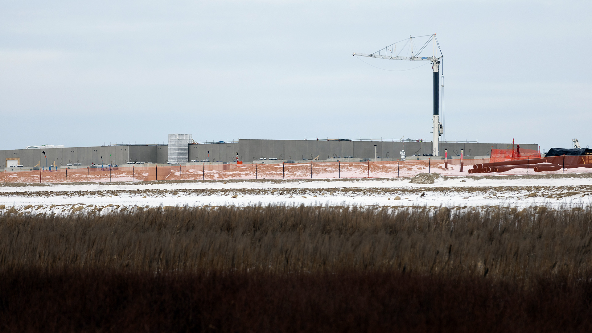 A construction crane stands next to concrete walls behind a mesh safety fence, with snow-covered fields and prairie grasses in the foreground, under an overcast sky.