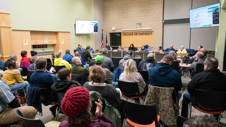 Multiple people sit behind a low legislative dais with multiple rows of people seated in rows of plastic and metal stacking chairs facing them, in a high-ceilinged room with the U.S. and Wisconsin flags in a corner next to a a door and a sign reading Village of DeForest, with two monitors mounted on walls and a digital clock reading 9:19:50 mounted in the middle of a row of cabinets. Multiple people sit behind a low legislative dais with multiple rows of people seated in rows of plastic and metal stacking chairs facing them, in a high-ceilinged room with the U.S. and Wisconsin flags in a corner next to a a door and a sign reading Village of DeForest, with two monitors mounted on walls and a digital clock reading 9:19:50 mounted in the middle of a row of cabinets.