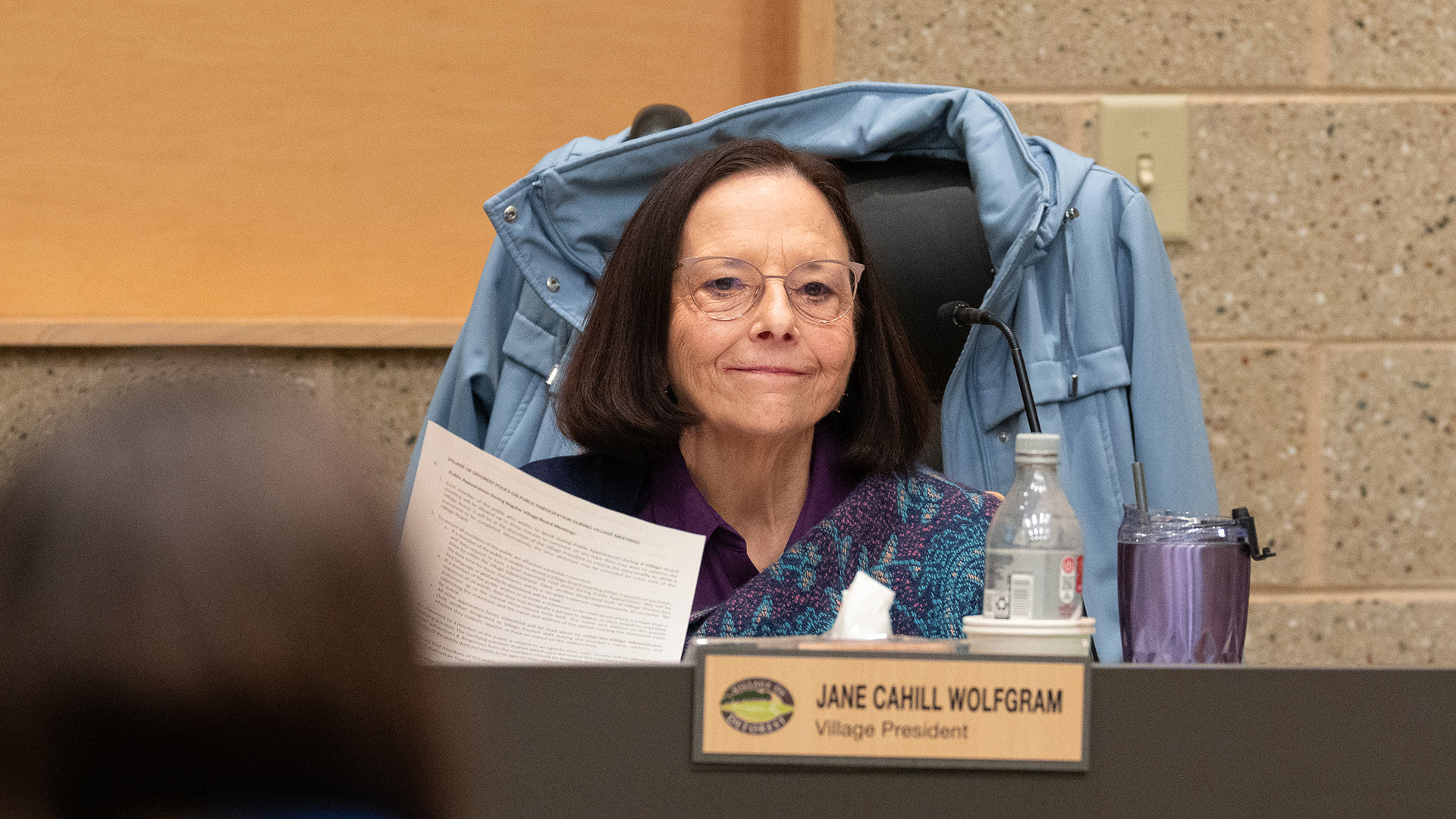 Jane Cahill Wolfgram holds a piece of paper with printed text in front of her while sitting in a high-backed leather chair with a coat draped over its back and behind a legislative dais with beverage containers on its surface and a name plate reading "Jane Cahill Wolfgram" and "Village President" affixed to its front, with a masonry wall in the background and an out-of-focus head of a seated person in the foreground.
