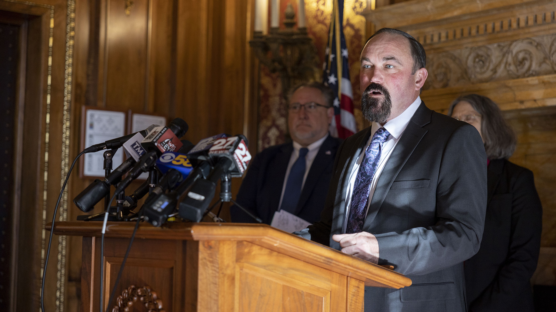 Clint Moses speaks into multiple microphones with the flags of different media organizations mounted to the top of a wood podium, with other people standing in the background of a room with a carved wood doorframe and fireplace mantel, toile wallpaper and U.S. flag.