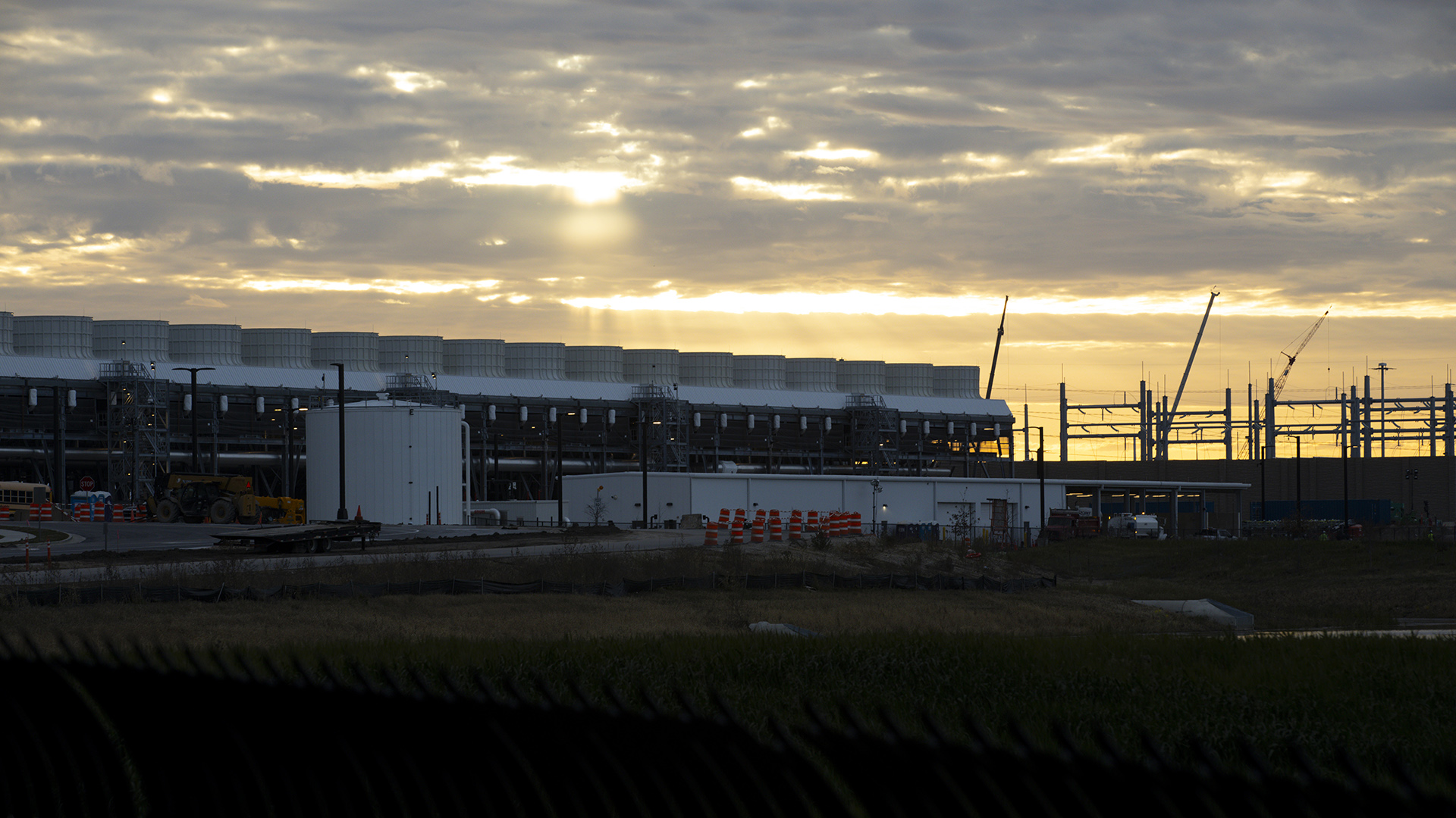 Sunlight filters through gaps in a mostly cloudy sky over a superstructure of metal supports and construction cranes and an adjacent large building with a row of cooling towers behind a bulk liquid storage tank and lower building next to a traffic-barrel lined roadway.