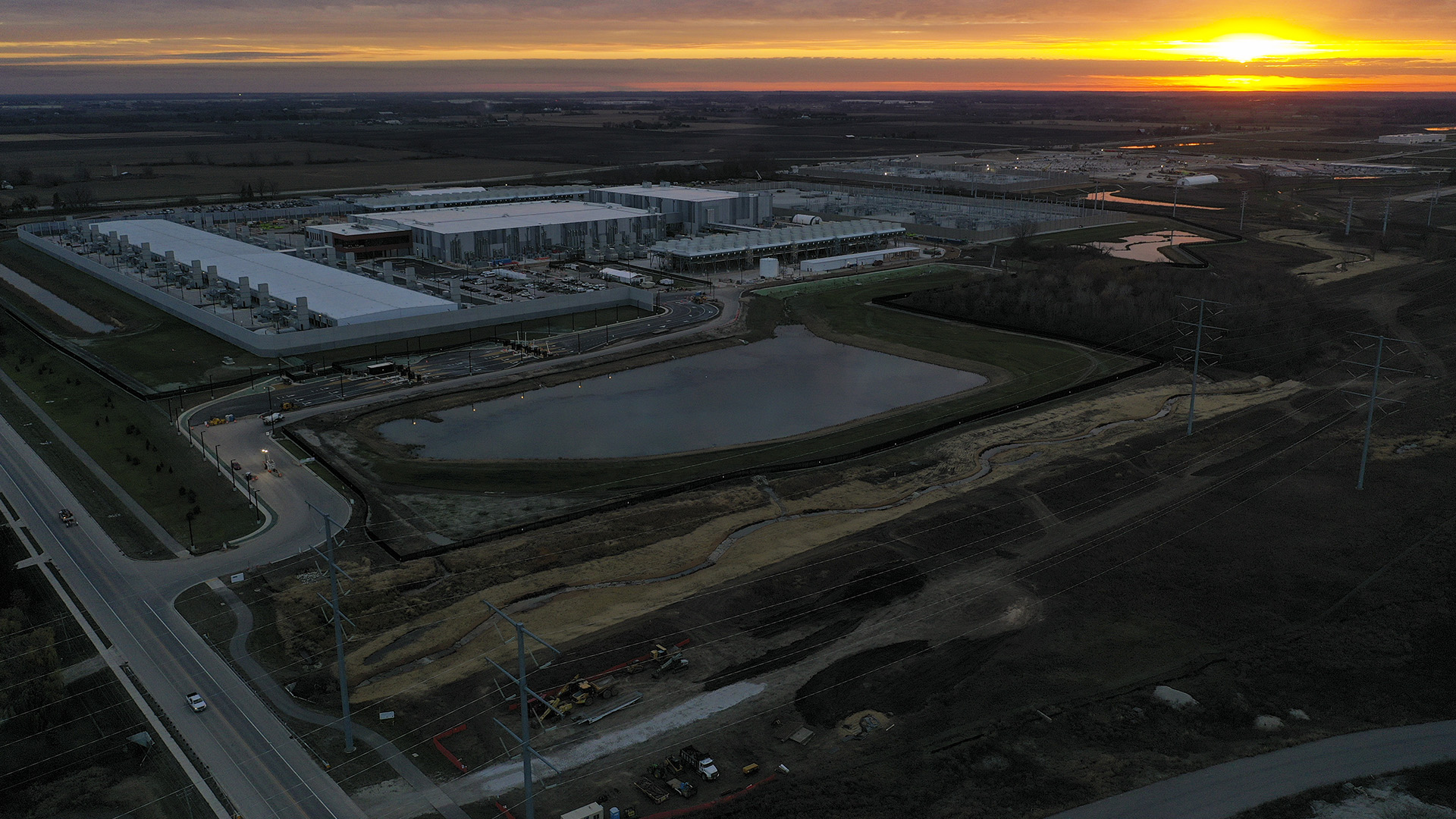 An aerial photo shows a retention pond, roadways, utility poles and lines, and a complex of multiple large buildings are illuminated by a sun setting in partly cloudy sky.