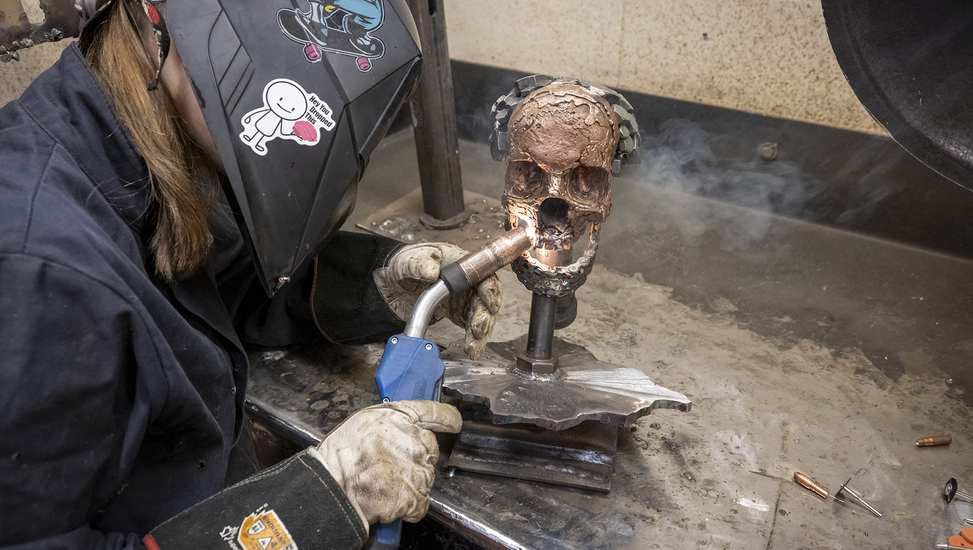 Izzy Chappell wears a welding mask welding gloves while using a torch on a metal sculpture in the shape of a human skull on top of a table.
