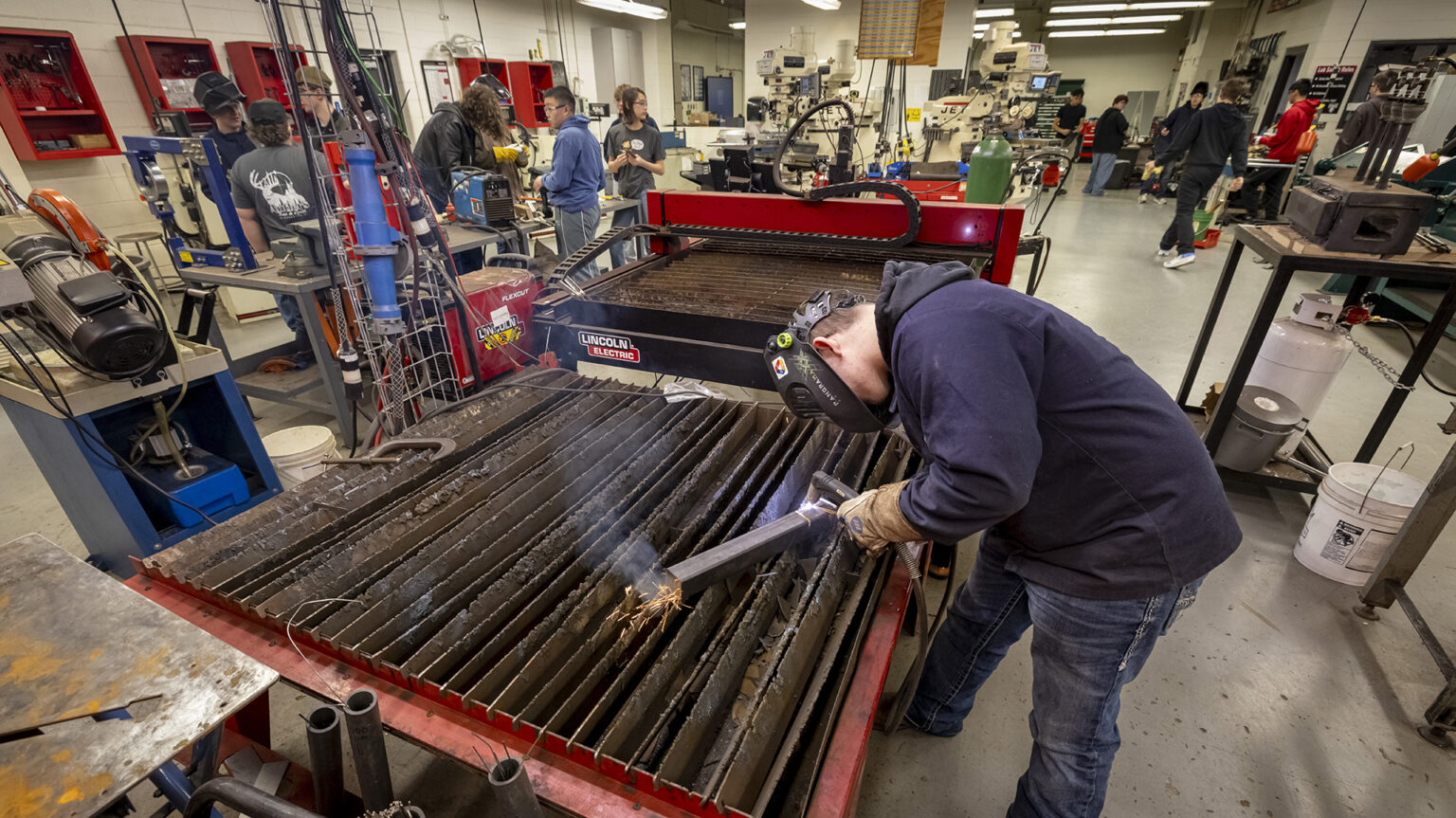 A student wearing a welding mask uses a welding torch to work on a piece of metal on top of a welding table in a large room with other manufacturing equipment on tables and in cases on walls, with other students standing and working on different projects in different locations. A student wearing a welding mask uses a welding torch to work on a piece of metal on top of a welding table in a large room with other manufacturing equipment on tables and in cases on walls, with other students standing and working on different projects in different locations.
