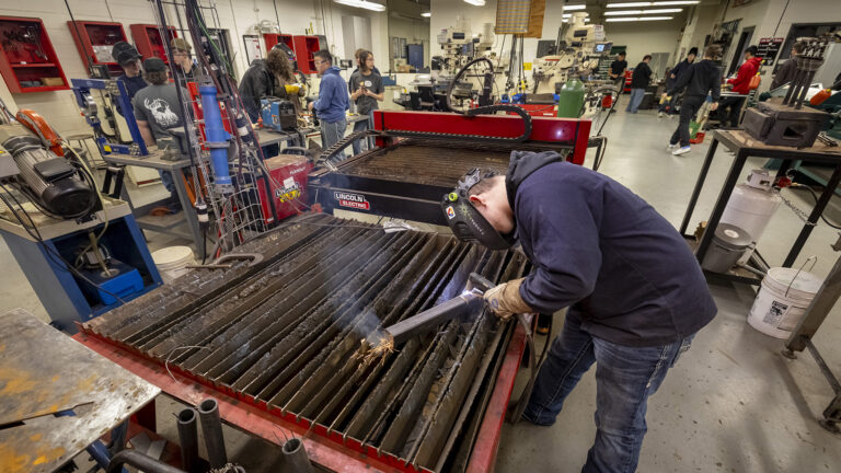 A student wearing a welding mask uses a welding torch to  work on a piece of metal on top of a welding table in a large room with other manufacturing equipment on tables and in cases on walls, with other students standing and working on different projects in different locations.