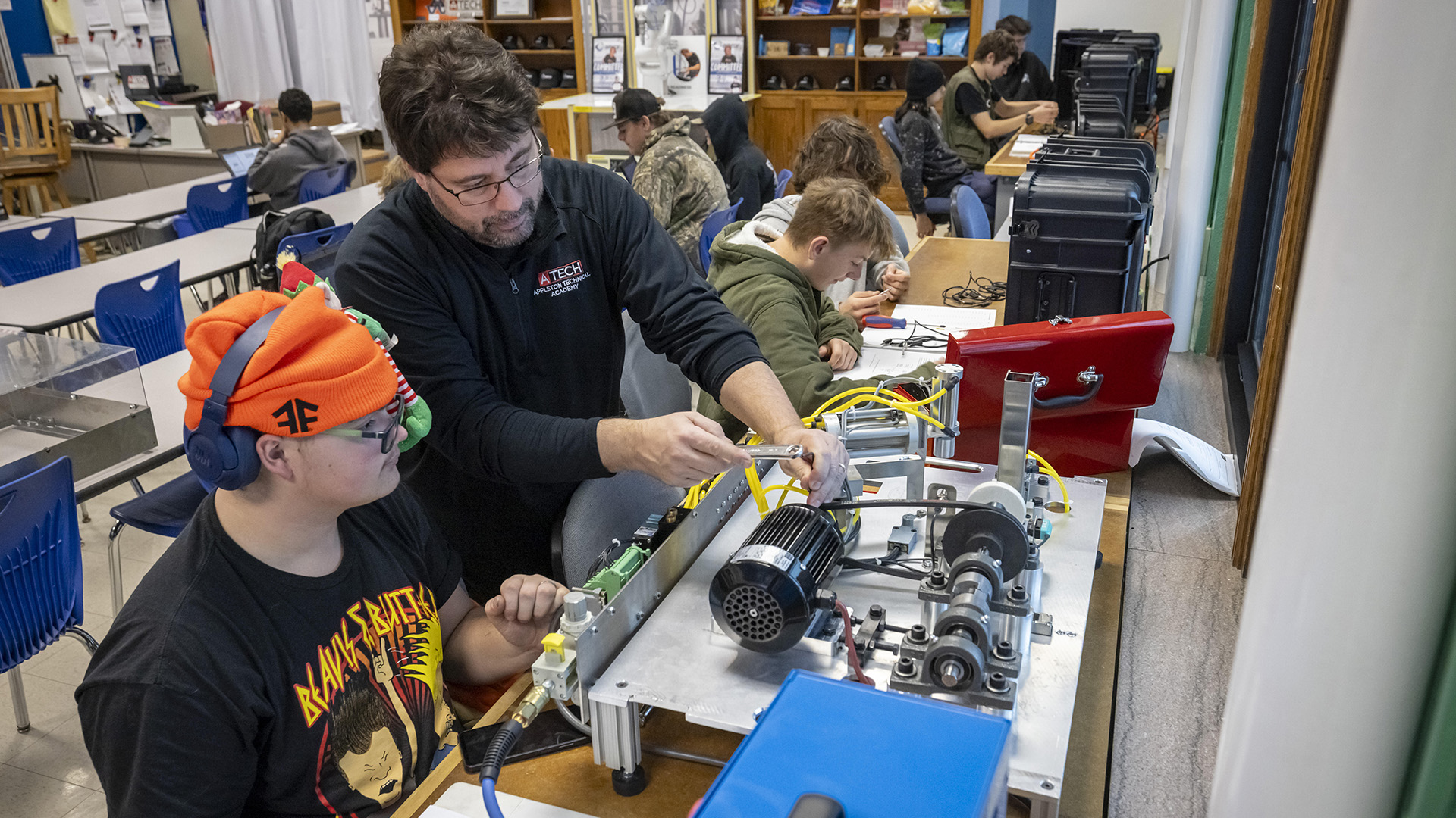 Loren Daane holds a wrench in his right hand and adjusts a piece of equipment on top of a table while standing next to a seated Joshua Bellman, with other students sitting at different tables in the background in a classroom.