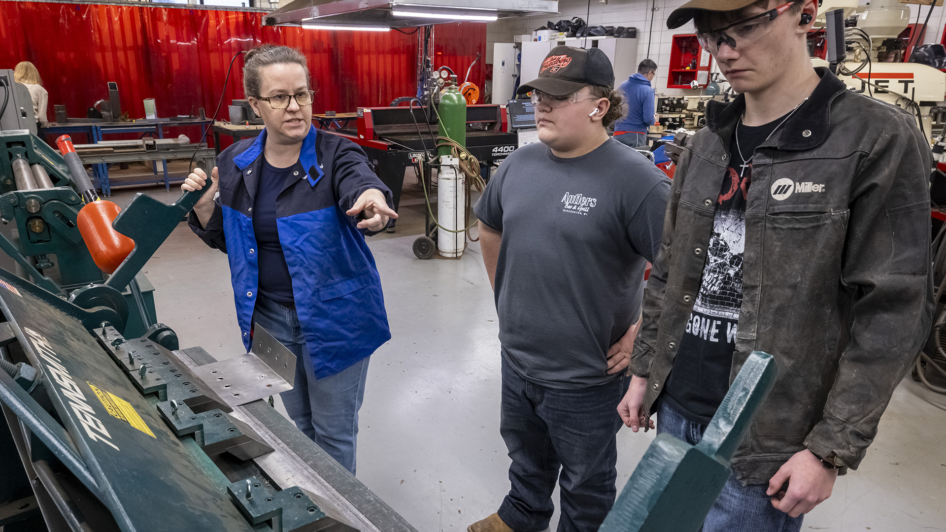 Carrie Giauque holds a lever attached to a large piece of manufacturing equipment in her right hand while pointing with the index finger of her left hand in front of two students standing next to her, in a room with other manufacturing equipment and people standing in the background.
