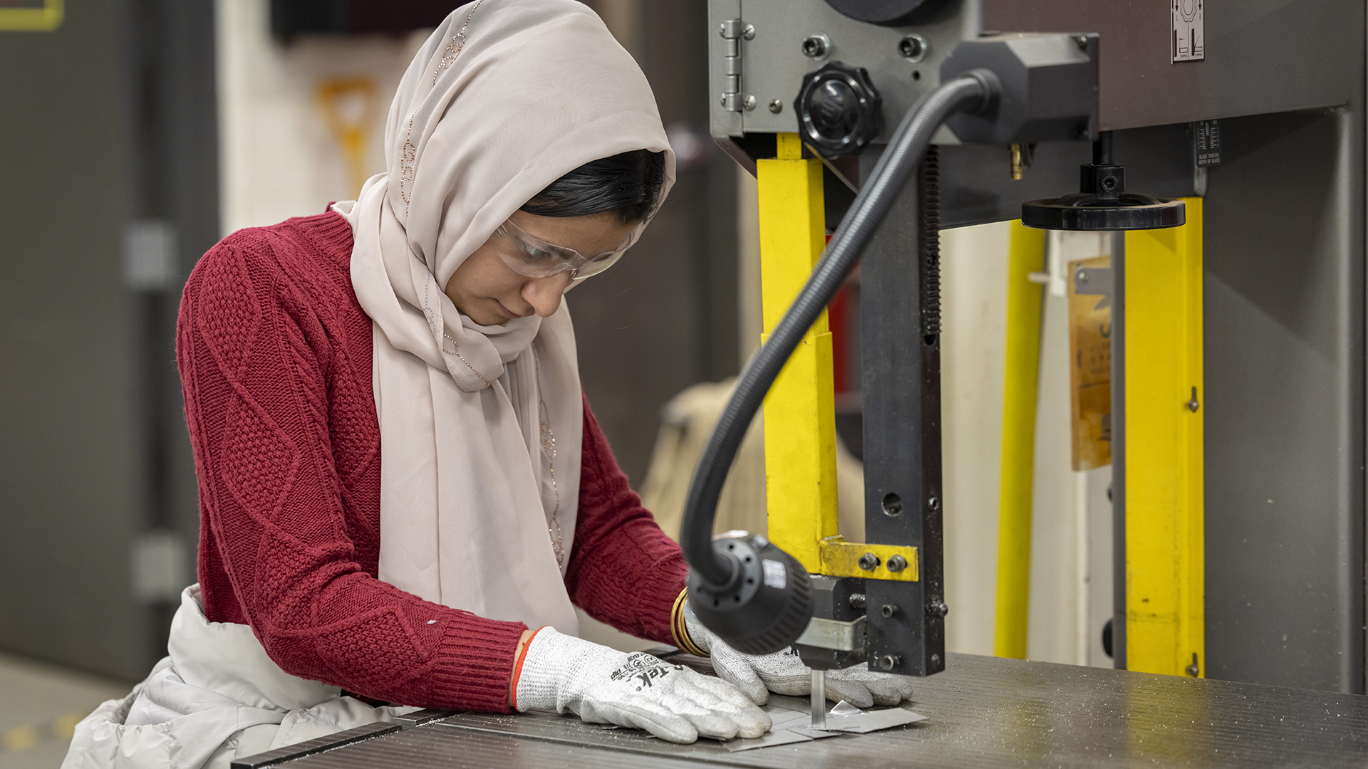 Marwa Nigravi wears safety gloves while using a bandsaw to cut a sheet of metal.