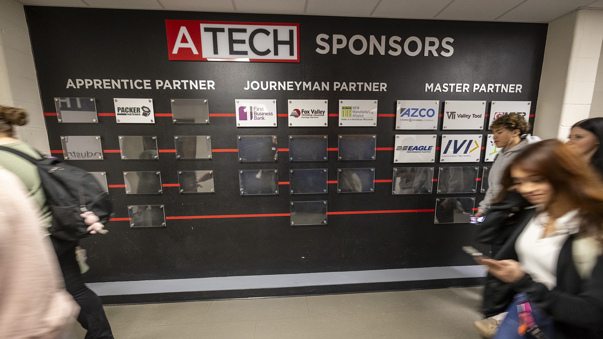 Out-of-focus students walk in a hallway in front of a wall with a sign reading "ATECH Sponsors" above the words "Apprentice Partner," "Journeyman Partner" and "Master Partner," with logos of different companies displayed in columns beneath each.