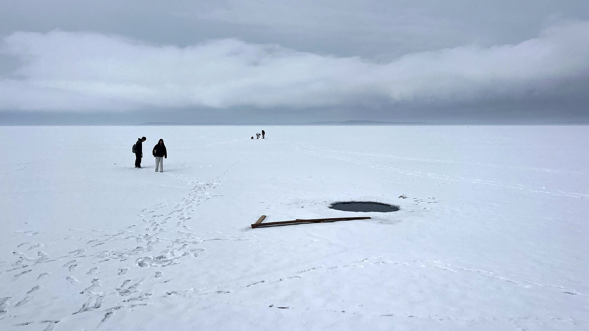 Two groups of multiple people walk on the snow-covered surface of a frozen lake past a circle of open water and under an overcast sky.