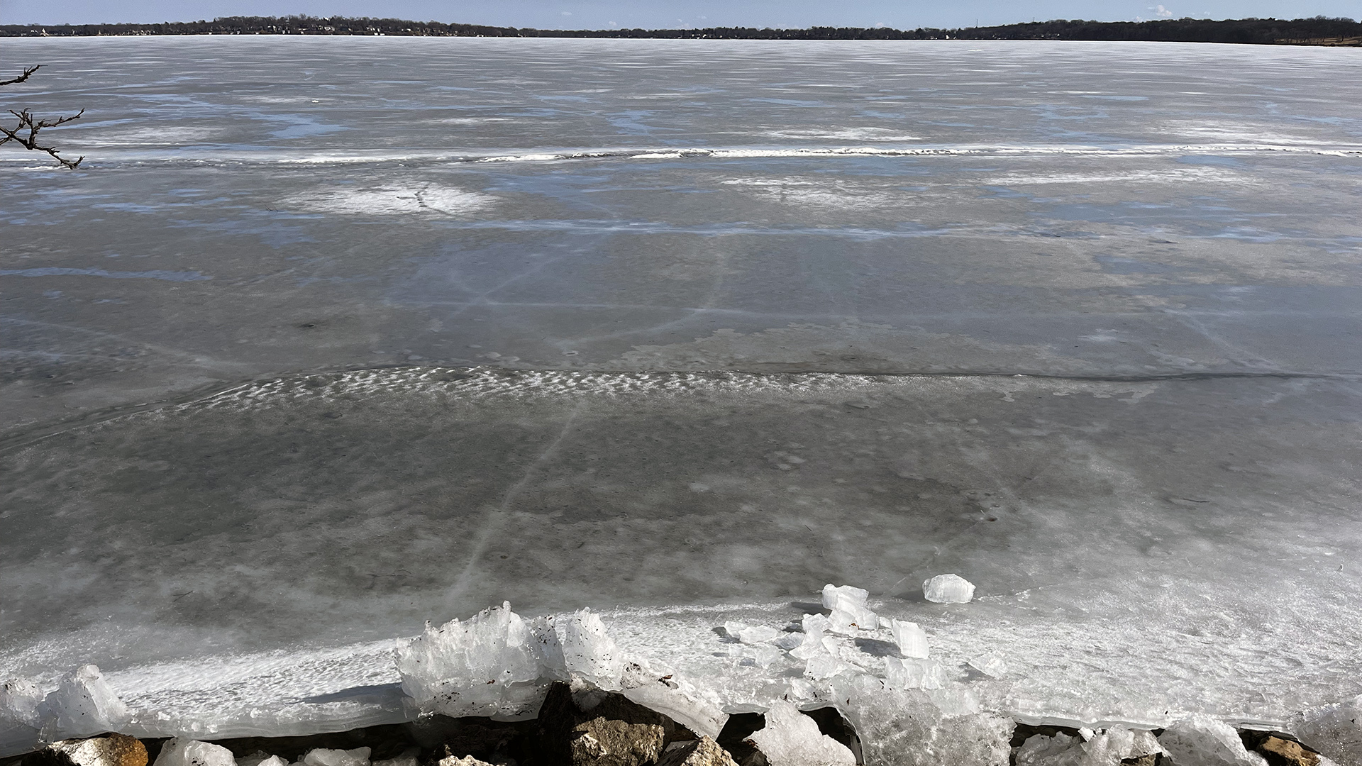 Puddles of water and patches of fragmented ice stand on the surface of a sheet of thin ice covering the surface of a lake, with a stone shore in the foreground and a tree-lined horizon on the far shore.