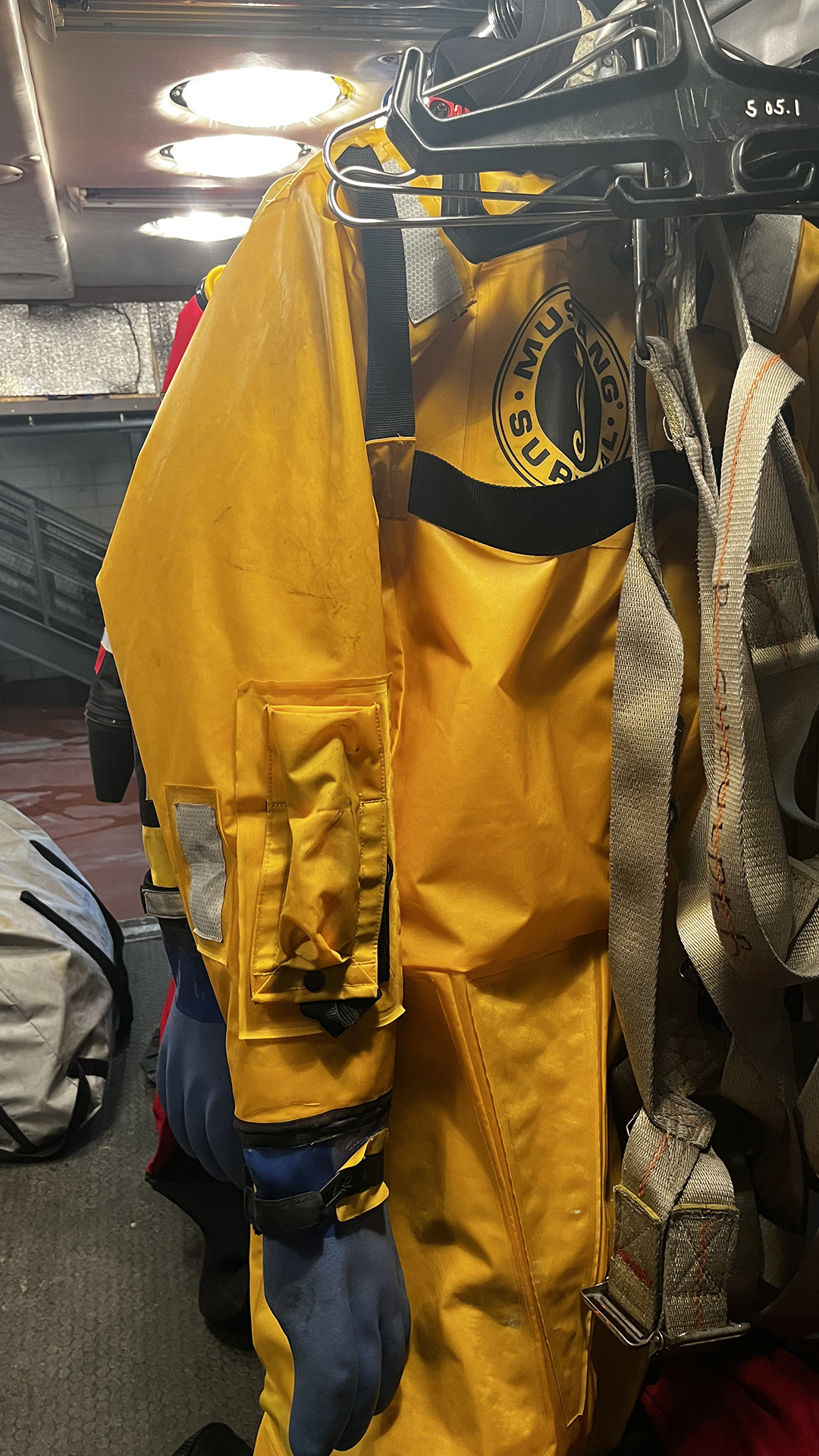 A survival suit and canvas harness are stored on hangers on a rack in a room with a duffel bag on the floor and multiple lights in the ceiling.