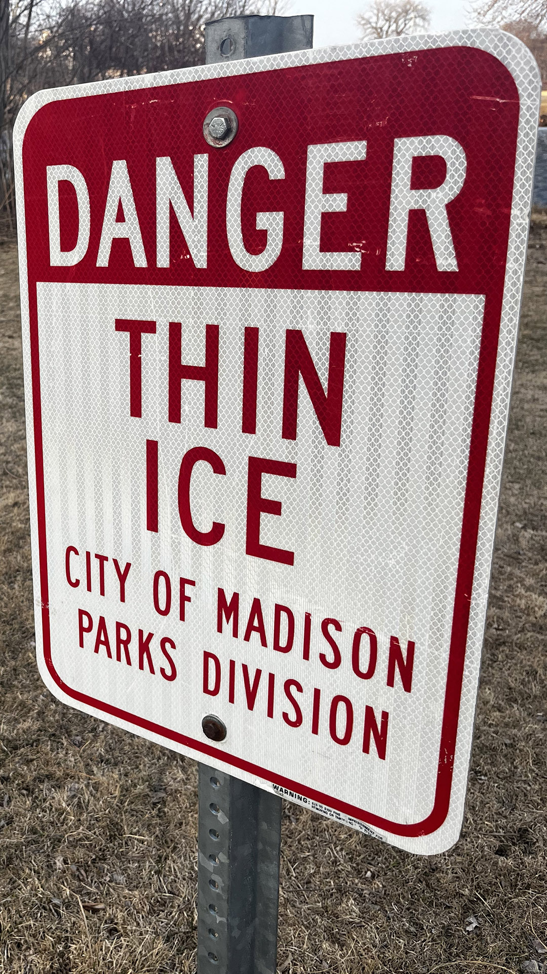 A metal sign with the words "Danger Thing Ice" and "City of Madison Parks Division" is displayed on a metal pole over a lawn with dormant grass and leafless trees in the background.