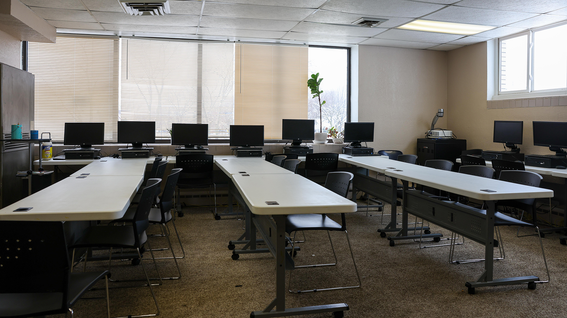 Three pairs of tables with plastic surfaces and metal legs on casters are arrayed in rows alongside stackable metal and plastic chairs in a room with multiple desktop computers with monitors arrayed on tables along two walls with windows with open and closed blinds.