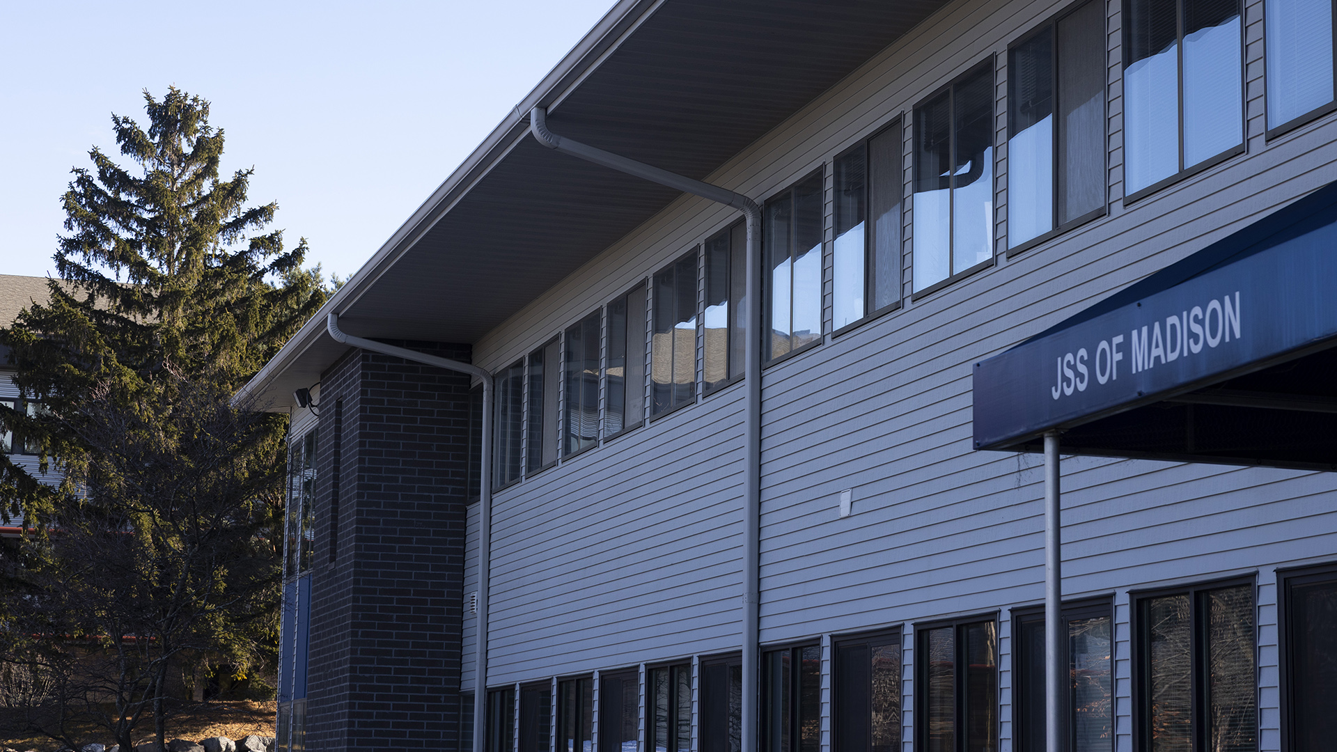 A two-story building with rows of windows on each floor and an awning with a sign on its side reading "JSS of Madison" stands in shadow in front of a coniferous tree and another building in the background illuminated by sunlight.