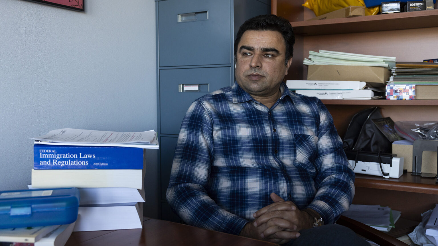 Zabi Sahibzada sits with his hands folded on his lap behind a desk with multiple books stacked on its surface and in front of a metal filing cabinet and a wood bookcase filled with file folders and other items on its shelves.