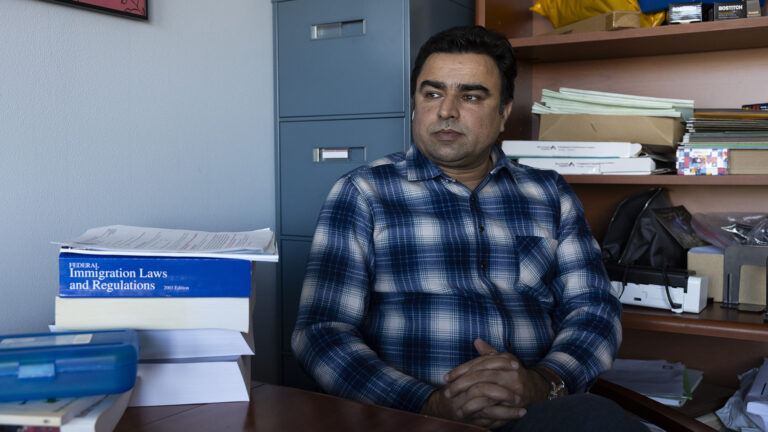 Zabi Sahibzada sits with his hands folded on his lap behind a desk with multiple books stacked on its surface and in front of a metal filing cabinet and a wood bookcase filled with file folders and other items on its shelves. Zabi Sahibzada sits with his hands folded on his lap behind a desk with multiple books stacked on its surface and in front of a metal filing cabinet and a wood bookcase filled with file folders and other items on its shelves.