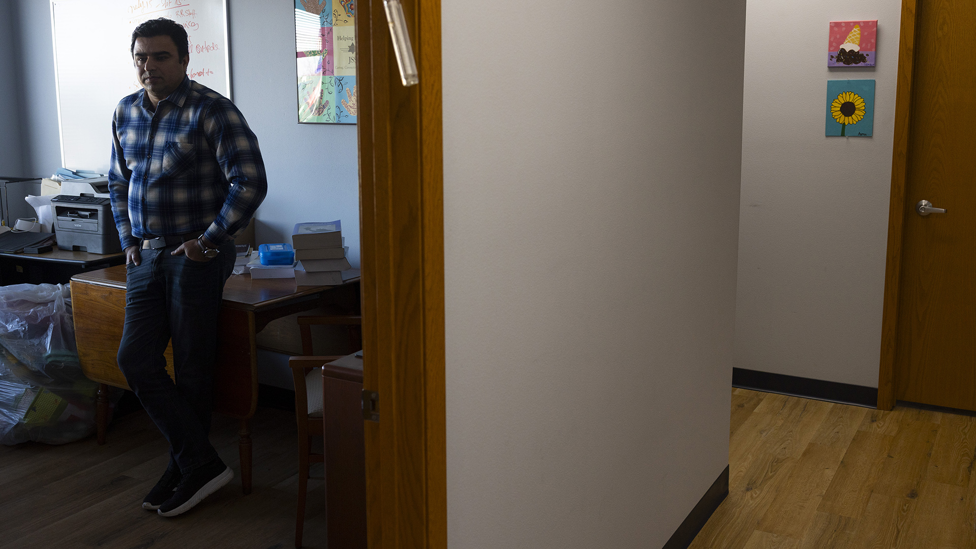 Zabi Sahibzada stands with his hands in his pants pockets while leaning against a wood desk in an office with a table with a printer on its surface and a whiteboard and framed poster displayed on a wall, while seen through a door facing a hallway with a wood floor leading to a closed door of another room.