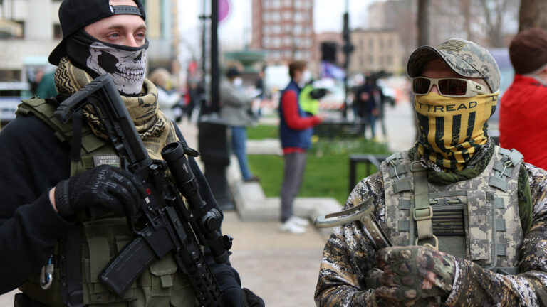 Two men wearing neck gaiters covering the lower half of their faces and carrying long guns stand on a sidewalk, with other out-of-focus people standing among park benches, light poles and buildings in the background.