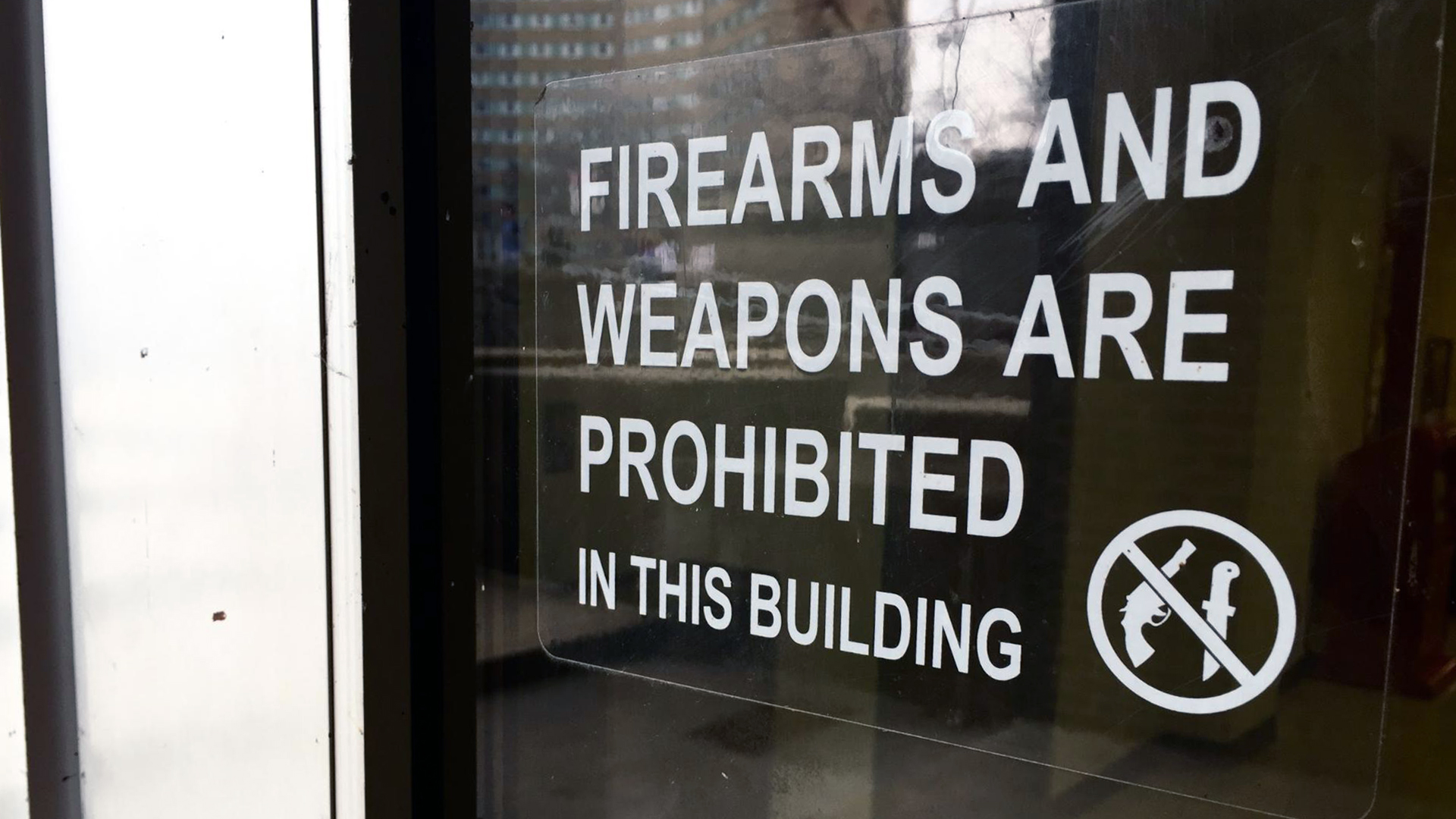 A plastic sign with the words "Firearms and Weapons Are Prohibited in this Building" next to a No symbol superimposed with graphics of a pistol and knife is mounted to a window, with a building and trees visible in a reflection.