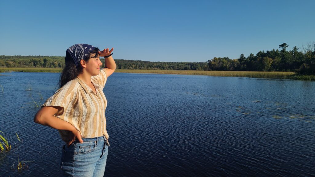 A young woman stands looking across a body of water, her hand shielding her eyes from the sun.