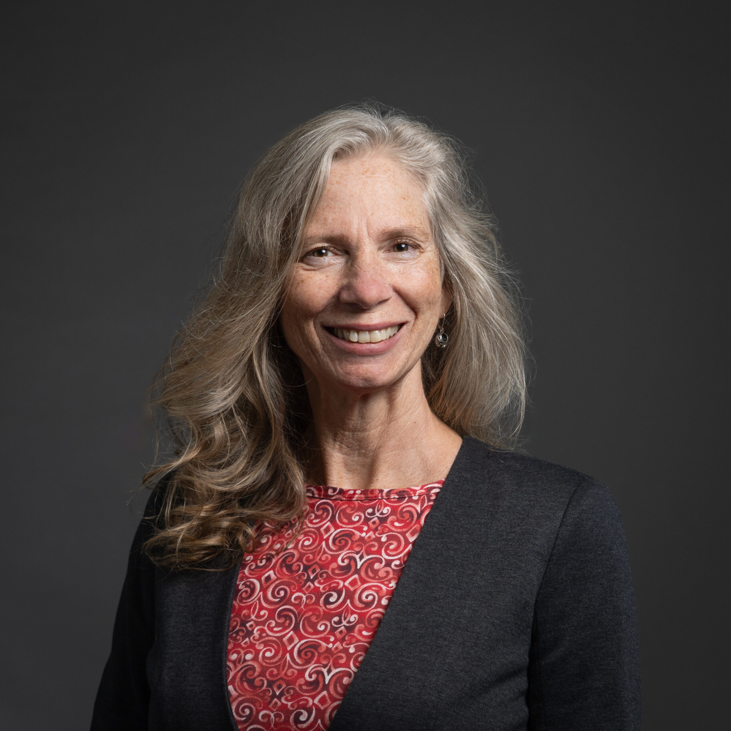 PBS Development Team Portrait Session 2025 - Sally Buffat Studio portrait of a woman with long gray hair, smiling, wearing a red patterned top and dark blazer against a dark backdrop.