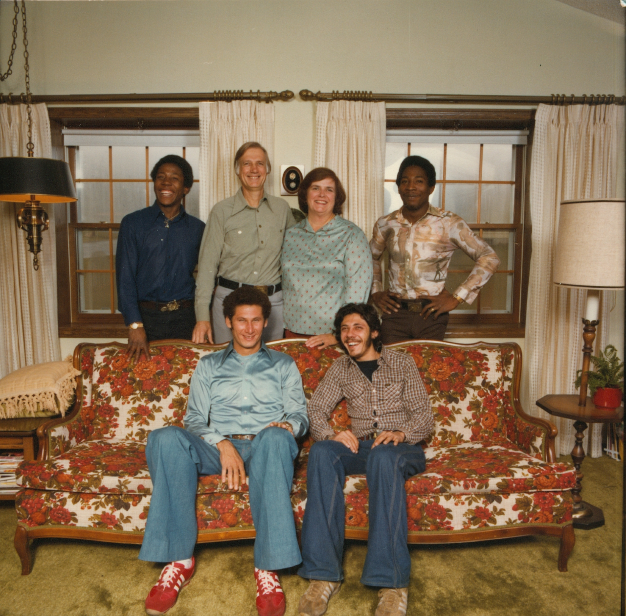 A group of six people smiling in a cozy living room. Three stand behind a floral couch, and three sit on it. The mood is warm and cheerful.