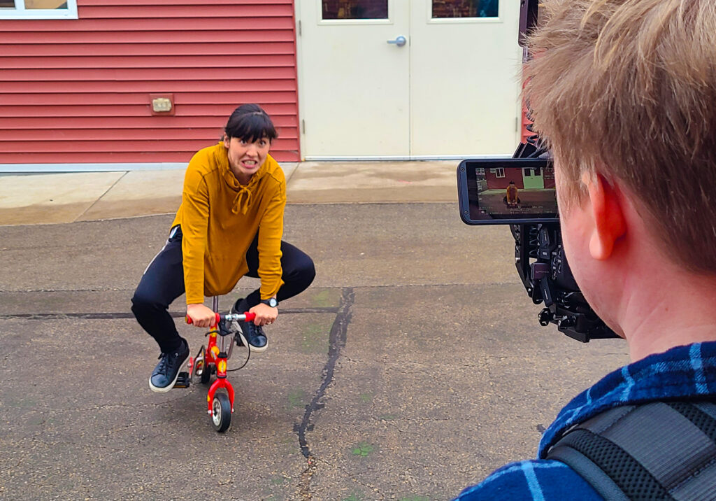 A young woman rides on a mini clown bike while being filmed for a video series.