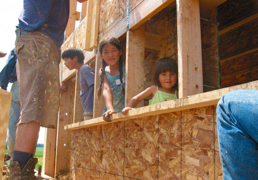 Three children peer out from the frame of a house. 