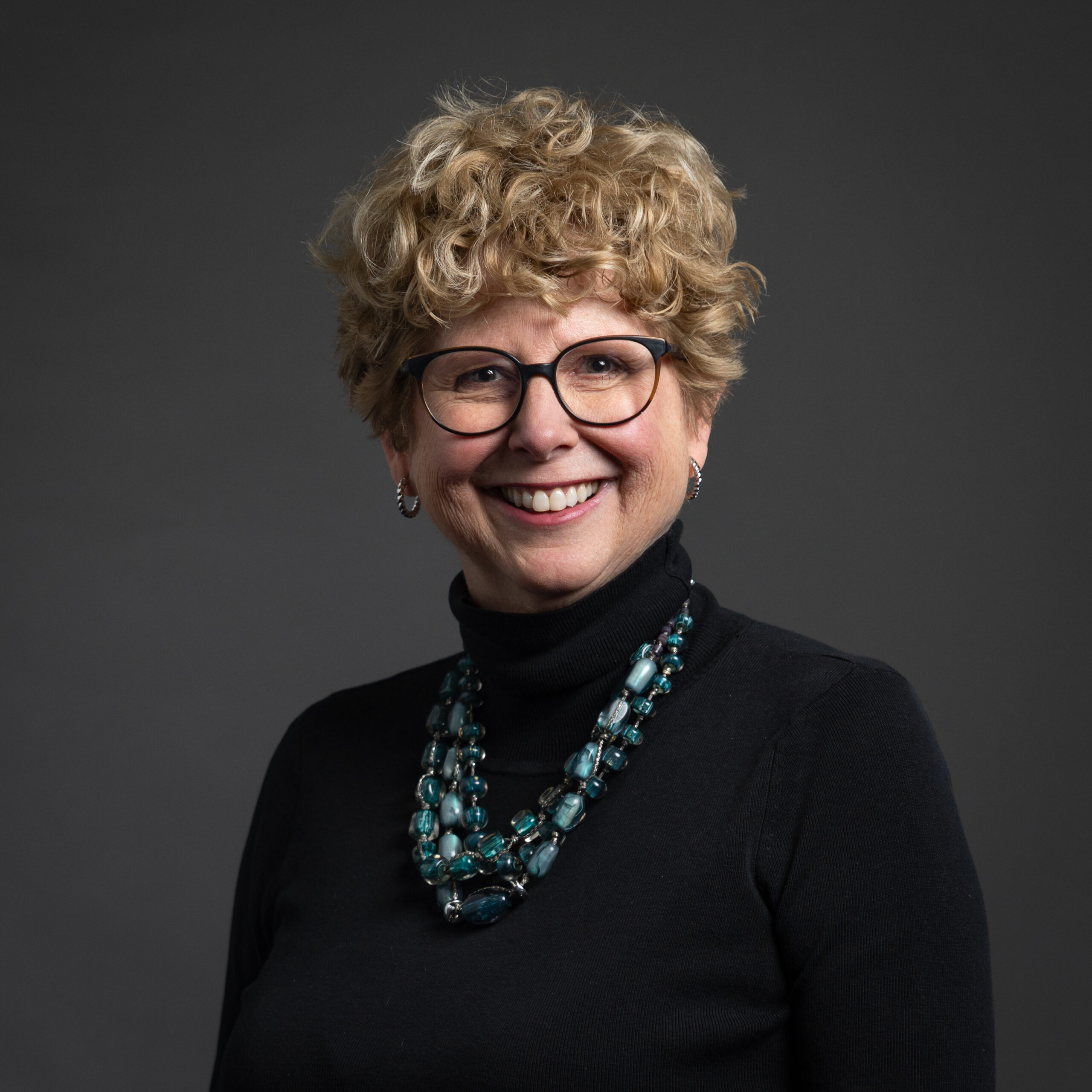 PBS Development Team Portrait Session 2025 - Lorrie Goodwin Professional headshot of a smiling woman with short, curly blond hair and round black glasses. She wears a black turtleneck sweater, silver hoop earrings, and a layered turquoise beaded necklace against a dark gray studio backdrop.