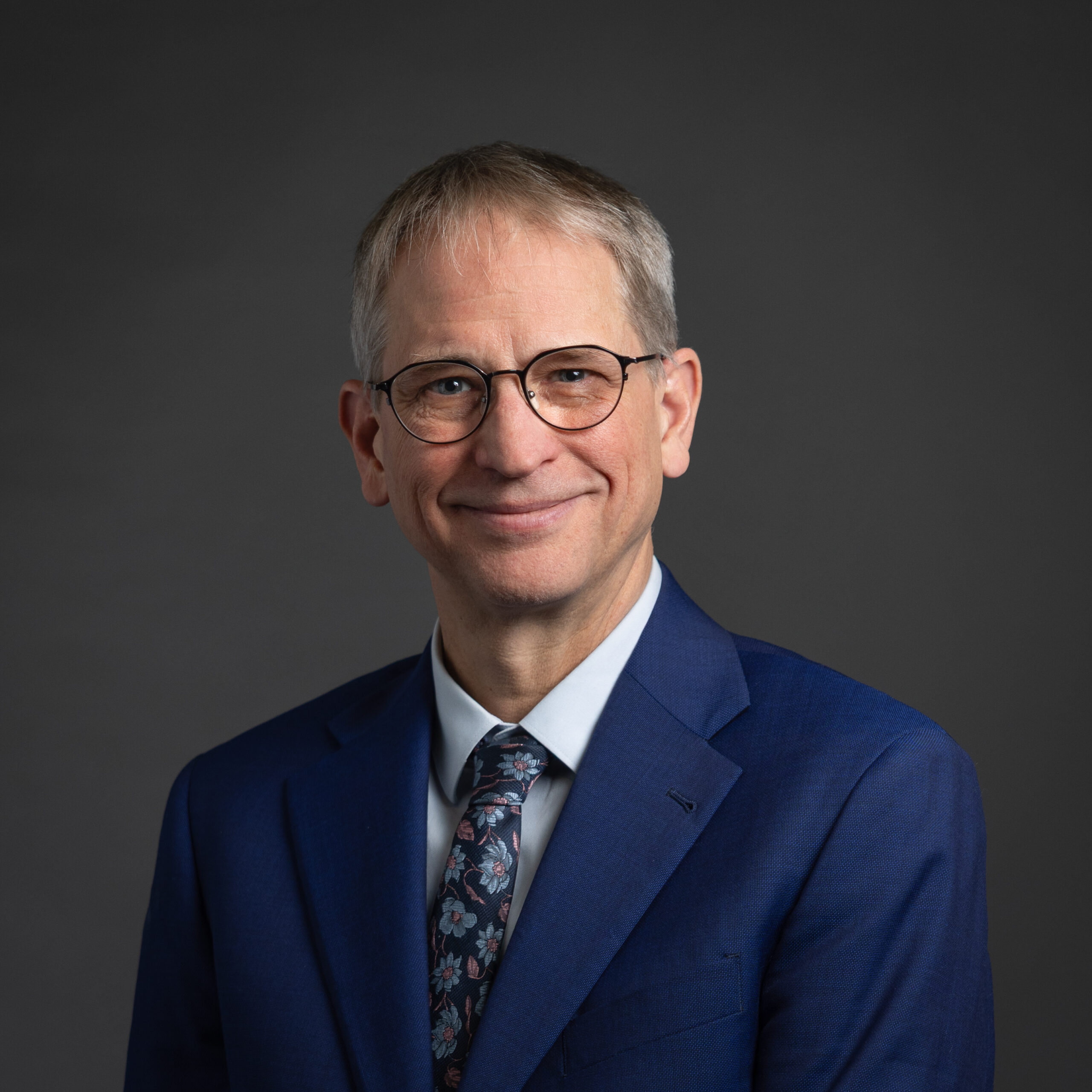PBS Development Team Portrait Session 2025 - Eric Greiling Professional headshot of a man with short gray hair and round glasses, wearing a blue suit and patterned tie against a dark gray studio backdrop.