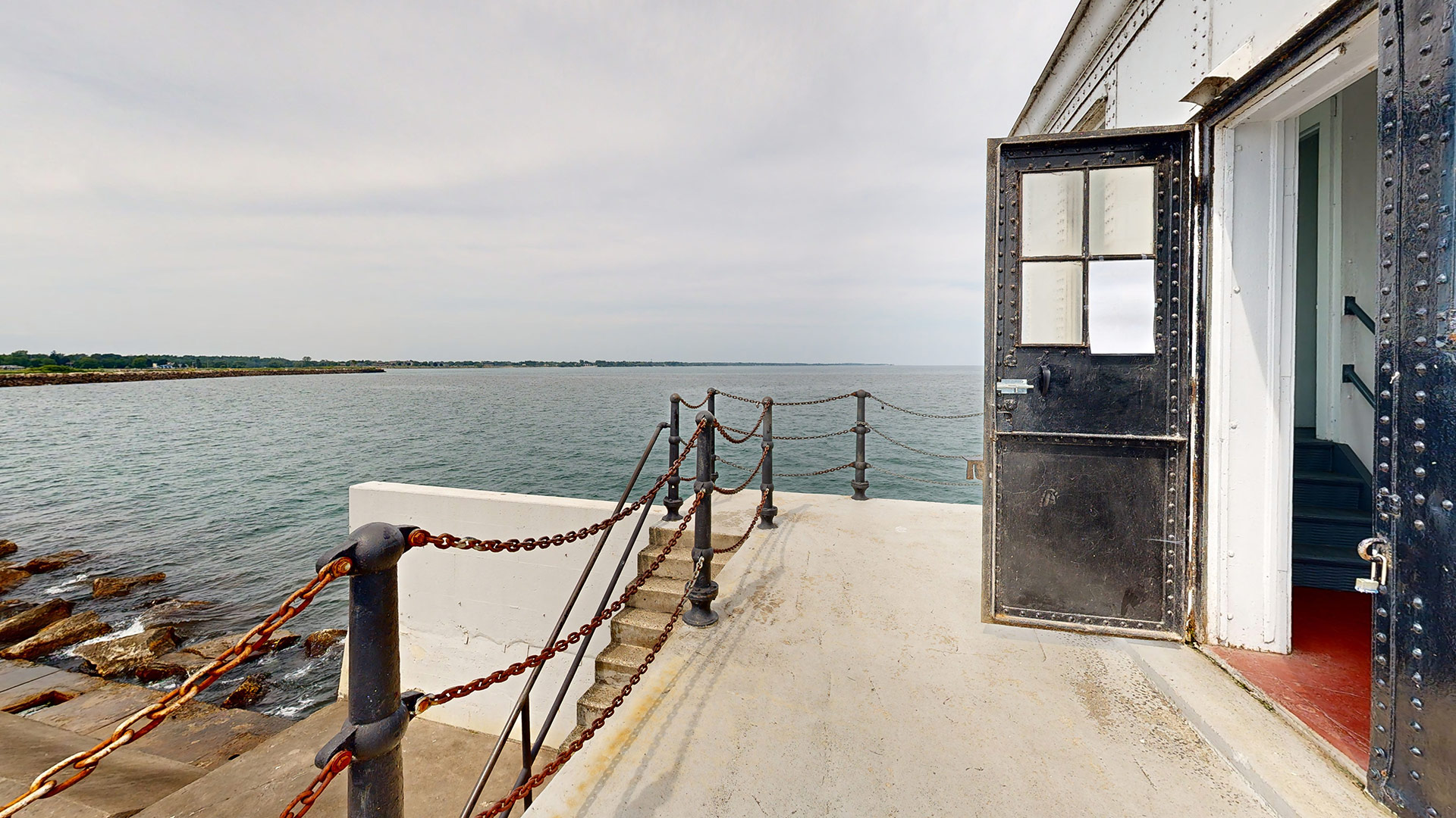 Open metal door of Manitowoc North Breakwater Lighthouse overlooking Lake Michigan and concrete pier with chain railing.