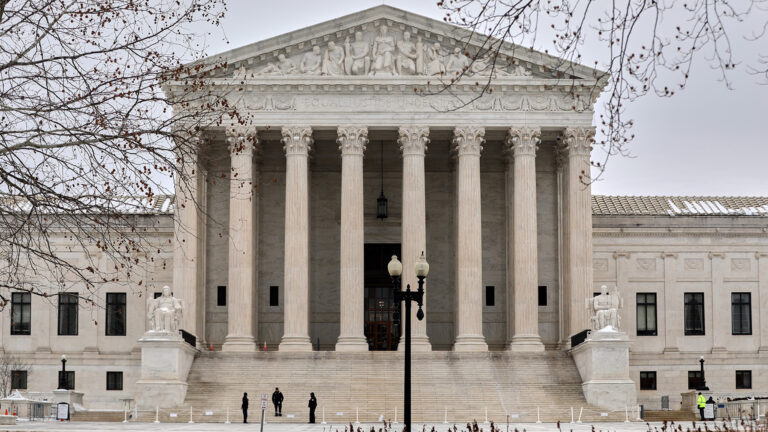 Several people stand on steps flanked by statues of two seated individuals outside an entrance to a multi-wing stone masonry building with a portico of columns upholding a pediment with sculptures of figures, with a street lamp, tree branches and grounds plants in the foreground. Several people stand on steps flanked by statues of two seated individuals outside an entrance to a multi-wing stone masonry building with a portico of columns upholding a pediment with sculptures of figures, with a street lamp, tree branches and grounds plants in the foreground.