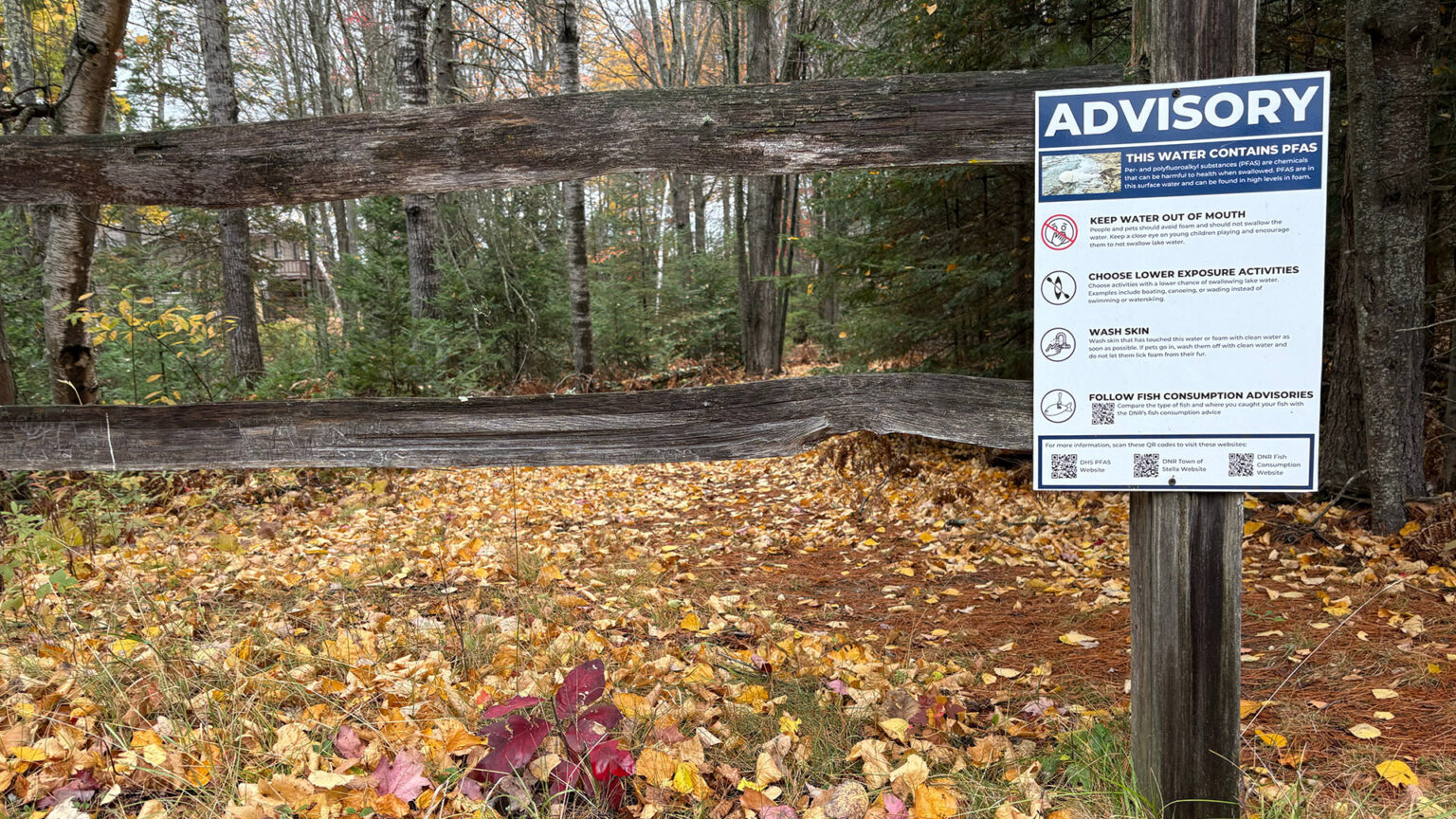 A sign with the title Advisory and lines reading This Water Contains PFAS, Keep Water Out of Mouth, Choose Lower Exposure Activities, Wash Skin and Follow Fish Consumption Advisories along with graphics, additional text and QR codes is attached to a post in a split-rail fence, with fallen leaves on the ground a building obscured by thick growth of trees in the background.