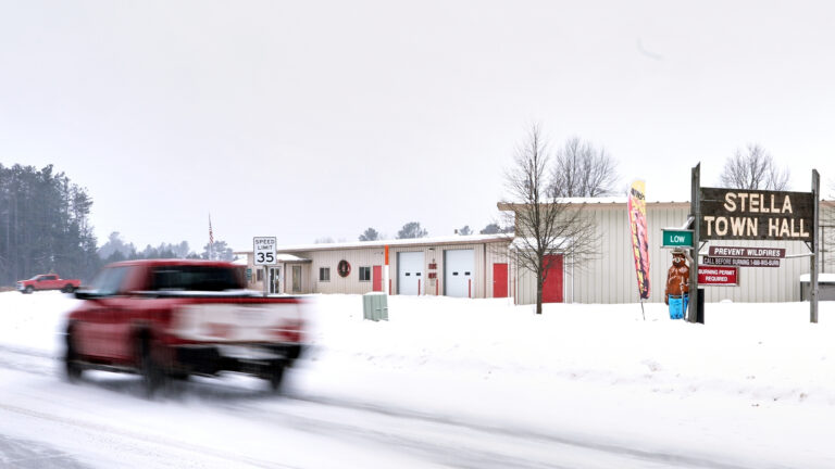A blurred pickup truck drives on a snow-covered road past a metal-sided building with an door at an entry vestibule, two garage doors and other doors along with a wood sign reading Stella Town Hall and a Smokey the Bear fire danger sign standing in snow-covered ground, with another tuck and trees in the background under an overcast sky.