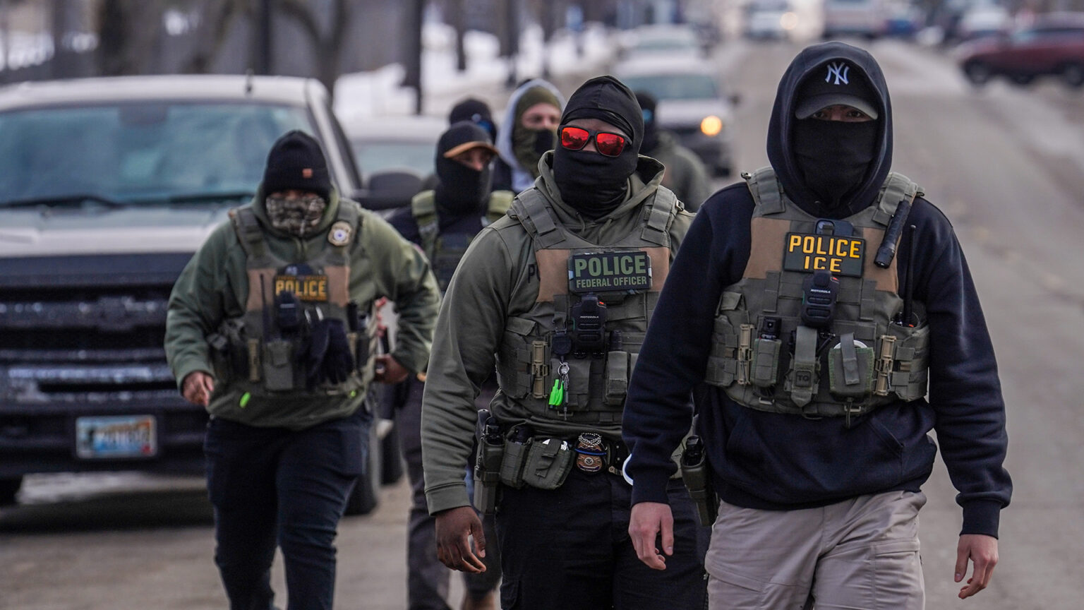 Multiple masked men wearing plate carrier vests with different removable patches reading Police ICE, Police Federal Officer and Police on top of different types and colors of apparel walk in a an uneven line down the middle of a street, with out-of-focus parked and driving vehicles in the background.